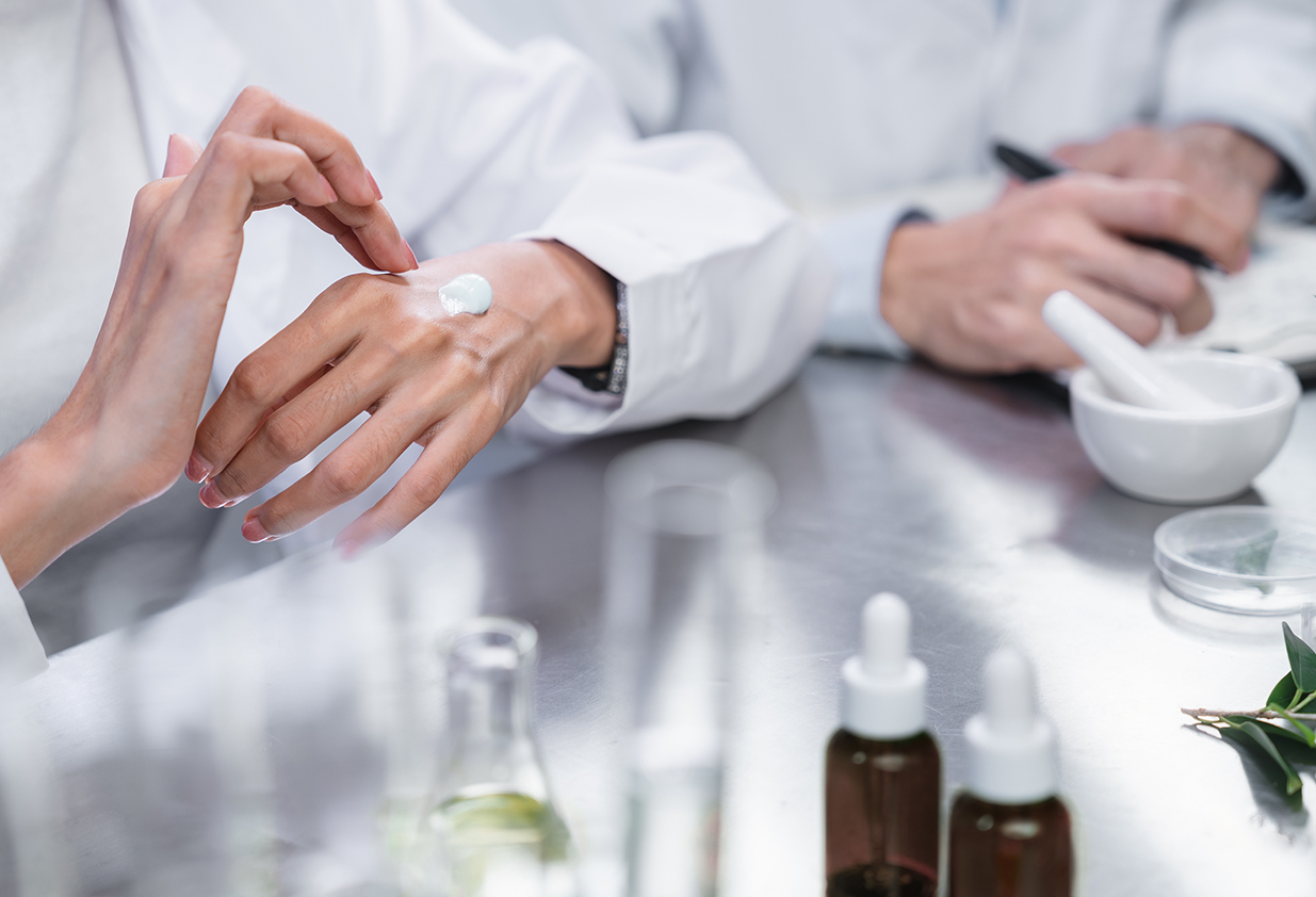 Two scientists at a table. One is holding a clipboard and the other is applying a cream to their hand as they review and discuss the formulation.